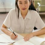 A focused young woman in a library writing in a notebook, symbolizing education and focus.