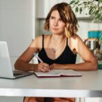 A woman in a black tank top writing with a laptop at a home desk during the day.
